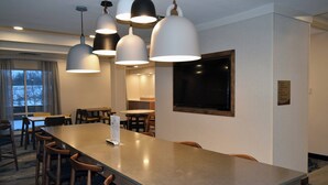 Un comedor de hotel con una larga mesa, varias sillas, iluminación colgante y vistas al exterior a través de una ventana.