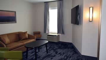Una habitación con un sofá marrón, una silla verde, una mesita pequeña, un televisor montado en la pared y una ventana con cortinas.