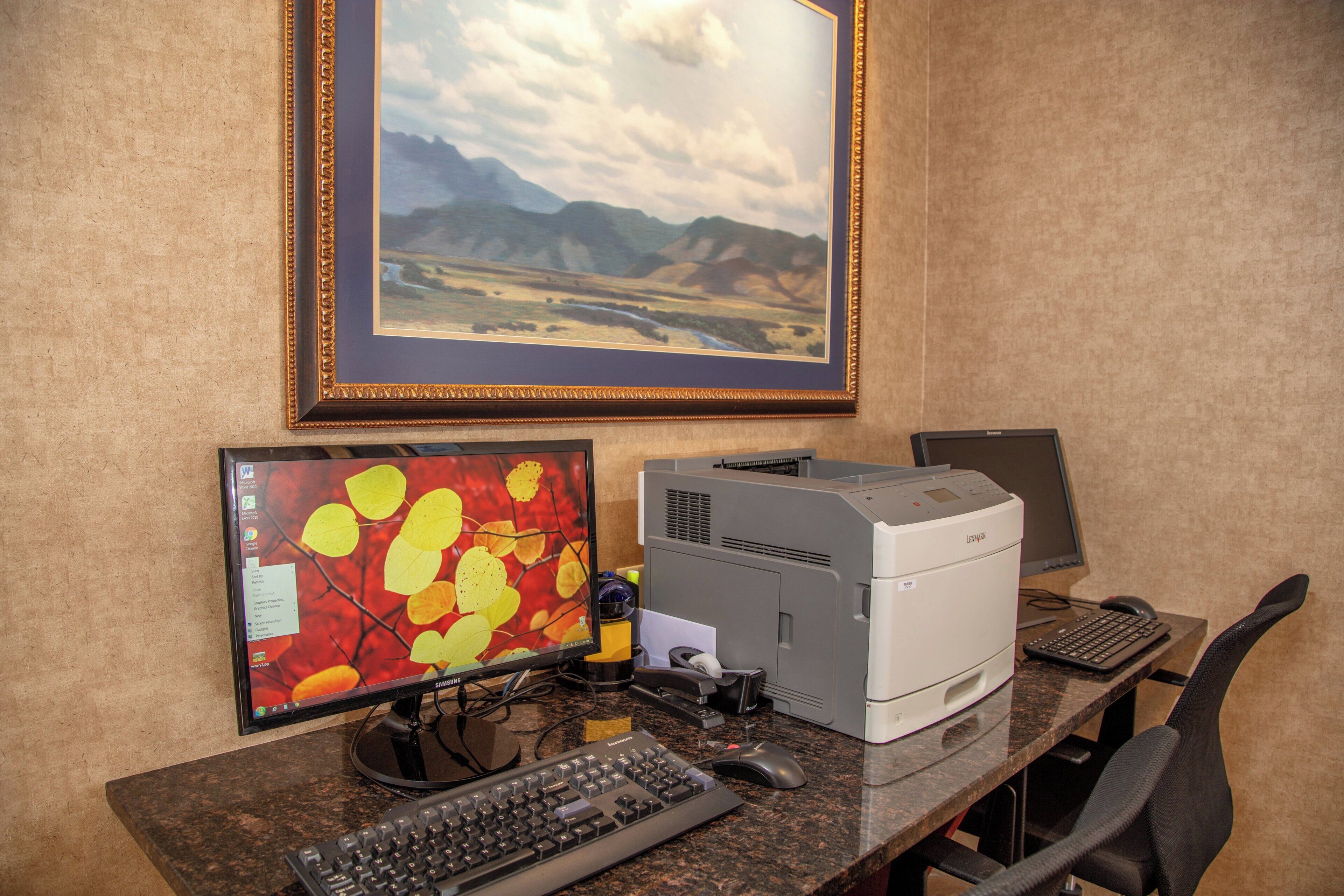 Un espace de travail équipé d’un ordinateur de bureau, d’un écran, d’une imprimante et d’un tableau représentant un paysage, accroché au mur.