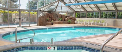 An indoor swimming pool with a glass ceiling, a rock feature, and seating area.