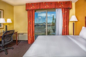 A hotel room with a large bed, a desk with two lamps, a chair, and a view of a building through a glass door. - Holiday Inn Express & Suites Richland by IHG (Richland)