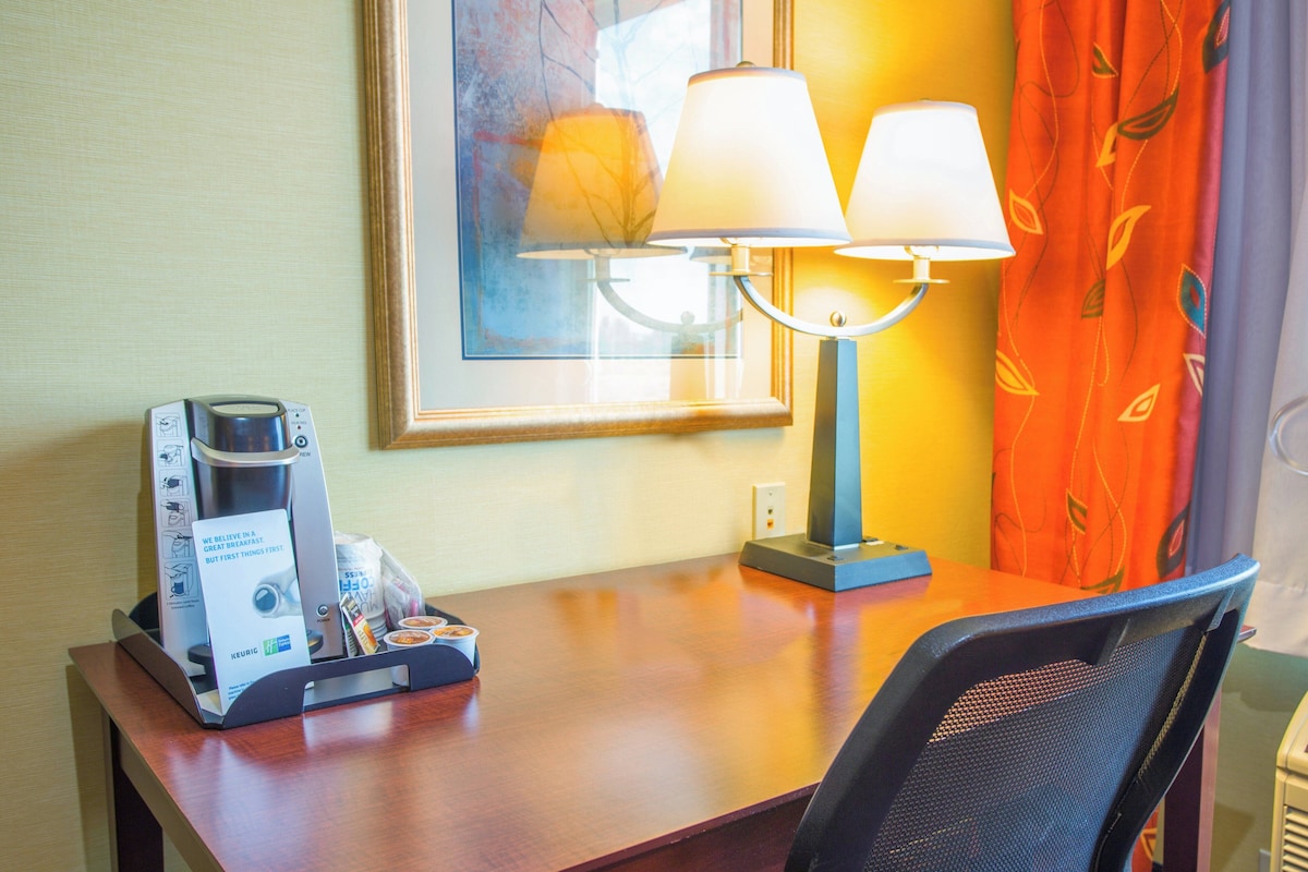 A hotel room with a wooden desk, a coffee maker, a lamp, and a framed picture on the wall.