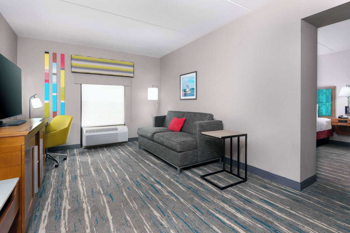 A modern hotel room with a grey sofa, a small table, a desk with a lamp, a colorful abstract painting, and a window with blinds.
