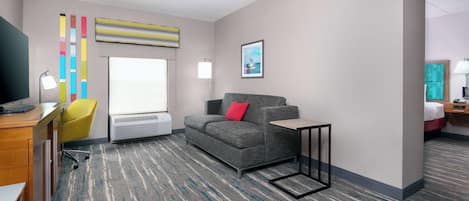A modern hotel room with a grey sofa, a small table, a desk with a lamp, a colorful abstract painting, and a window with blinds.