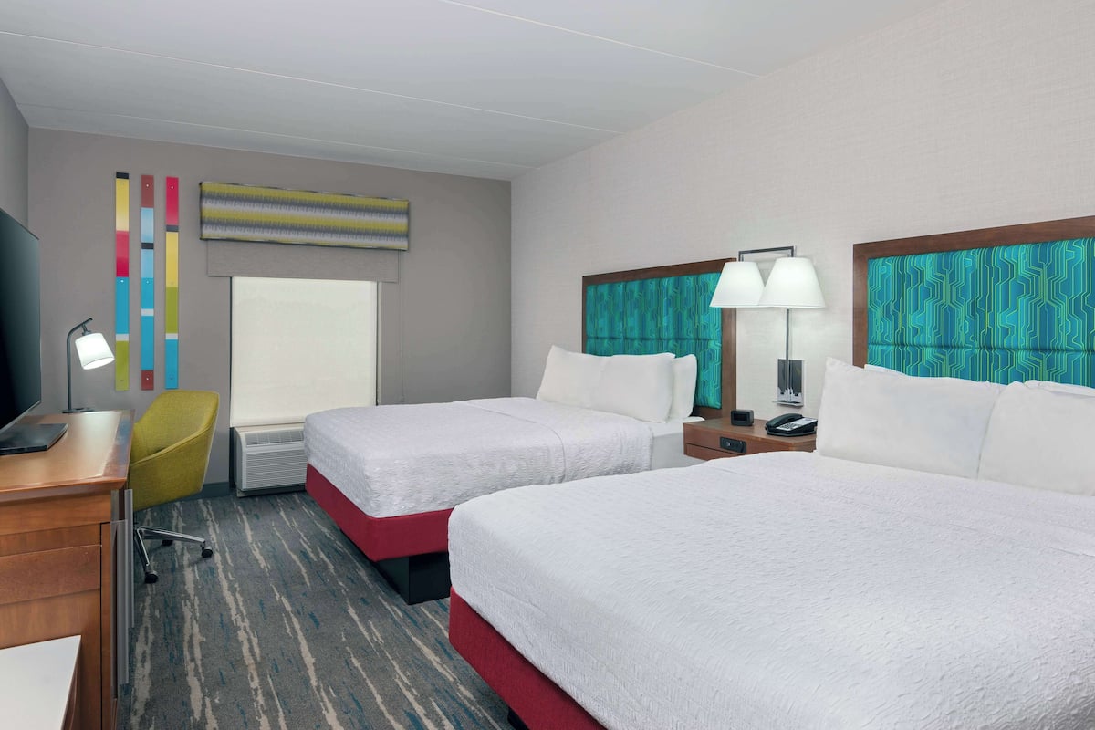 A hotel room with two beds, a desk with a computer, a chair, a window, and a wall with colorful decor.