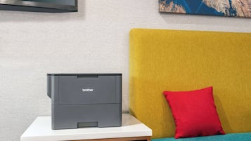 Una impresora Brother sobre una mesa blanca, junto a un sofá amarillo con un cojín rojo.
