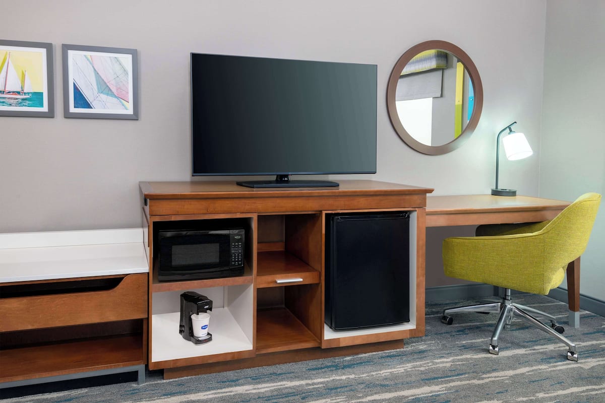 A hotel room with a wooden desk, a flat-screen TV, a microwave, a mini-fridge, a yellow chair, and a round mirror.