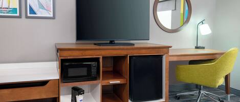 A hotel room with a wooden desk, a flat-screen TV, a microwave, a mini-fridge, a yellow chair, and a round mirror.