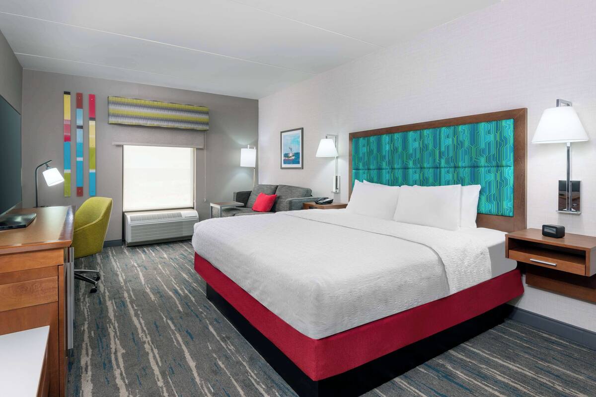A hotel room with a large bed, a desk, a chair, a window, and a wall with a painting.