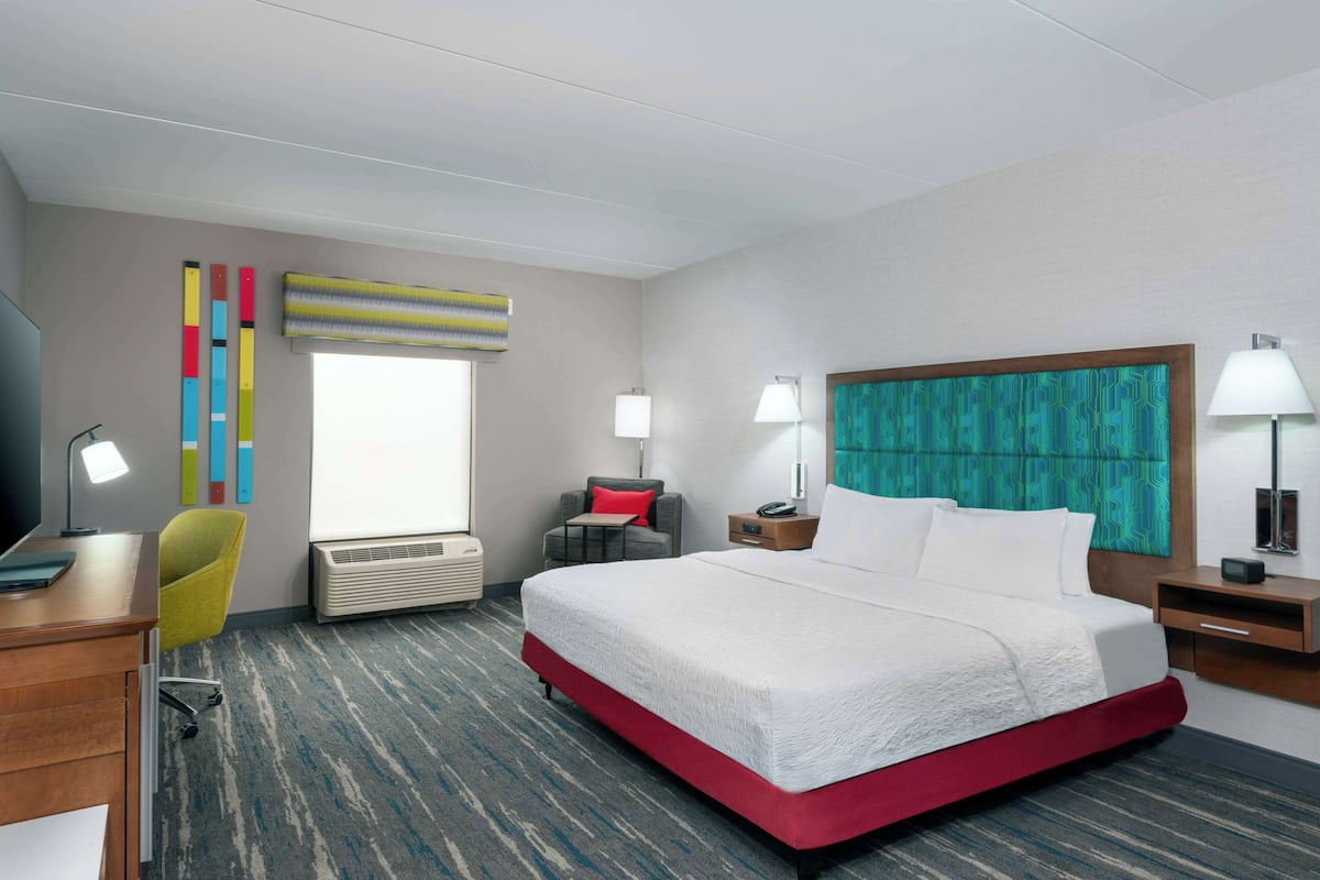A hotel room with a large bed, a desk with a computer, a chair, a desk lamp, a nightstand with a lamp, and a window with blinds.