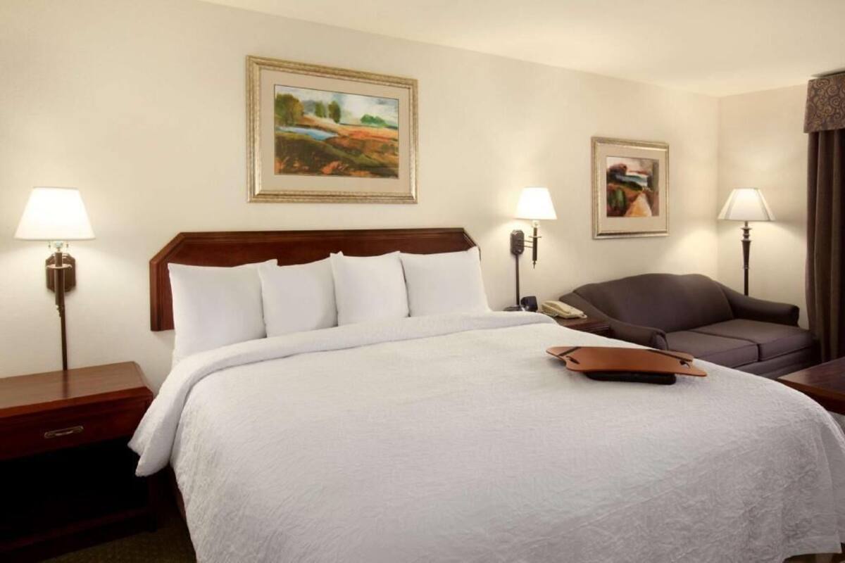 A hotel room with a large bed, two bedside lamps, a brown leather sofa, and two framed paintings on the wall.