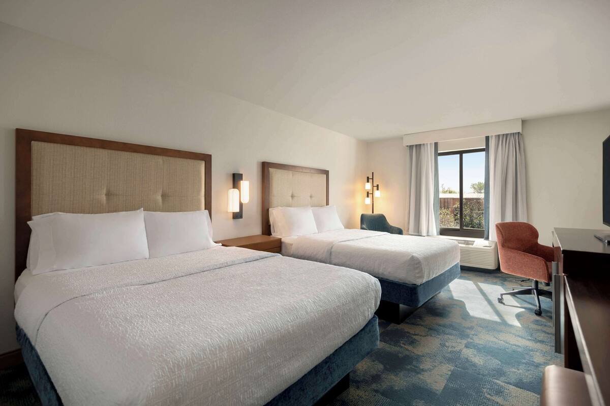 A hotel room with two beds, a desk, a chair, and a large window with curtains.