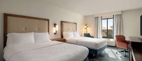 Habitación de hotel con dos camas, un escritorio, una silla y un amplio ventanal con cortinas.