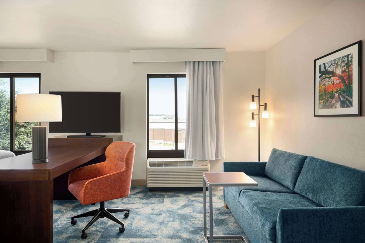 A hotel room with a desk, an orange office chair, a blue sofa, a TV, an air conditioner, and a window with curtains.