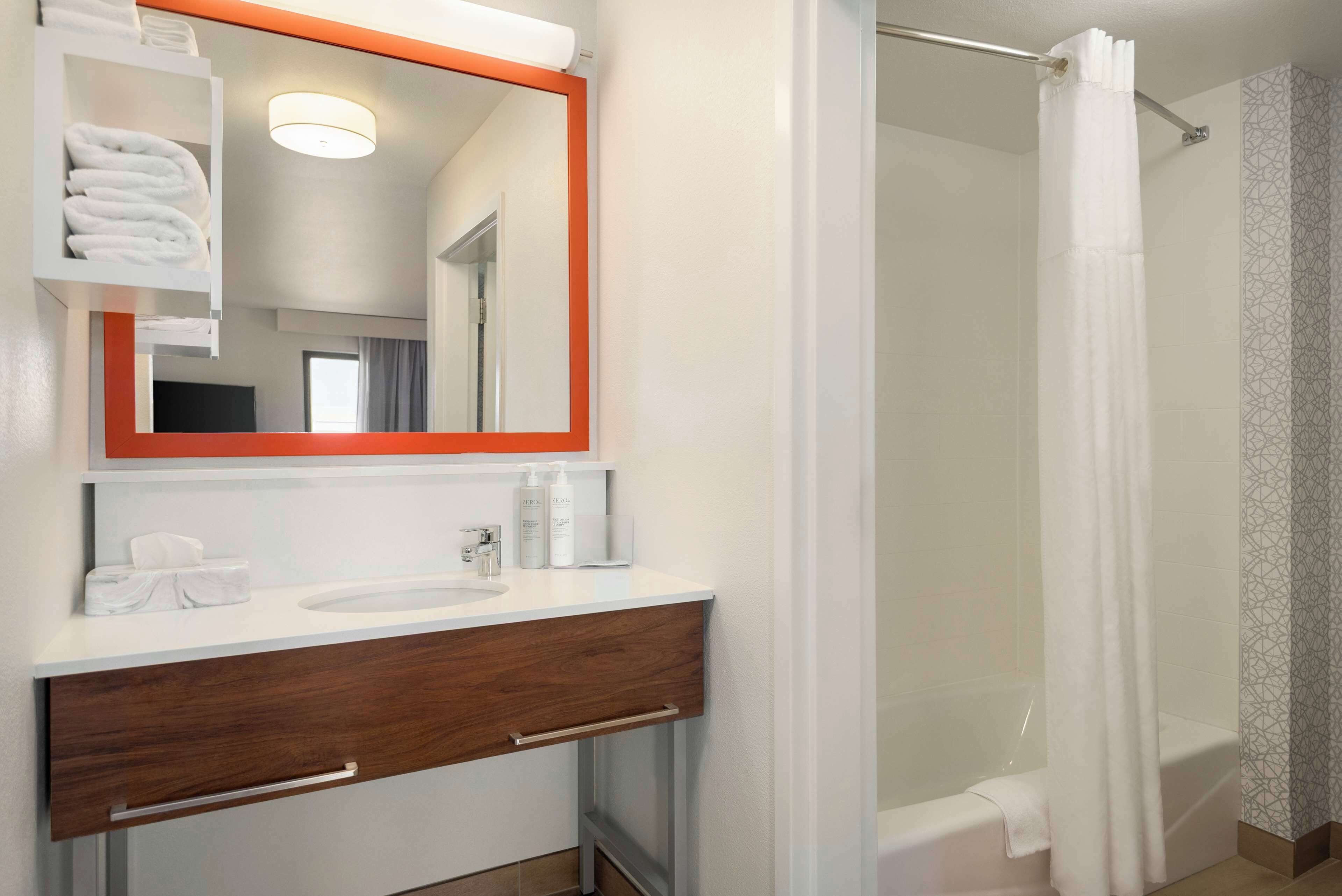 Une salle de bain moderne avec un meuble-lavabo blanc, un grand miroir avec un cadre orange, une douche avec un rideau blanc et un porte-serviettes avec des serviettes pliées.
