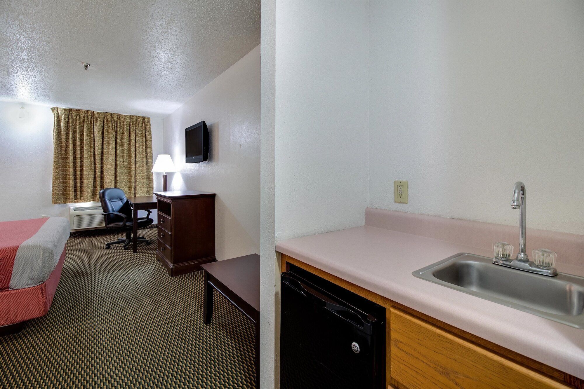 A hotel room with a bed, desk, chair, lamp, TV, and a kitchenette with a sink.