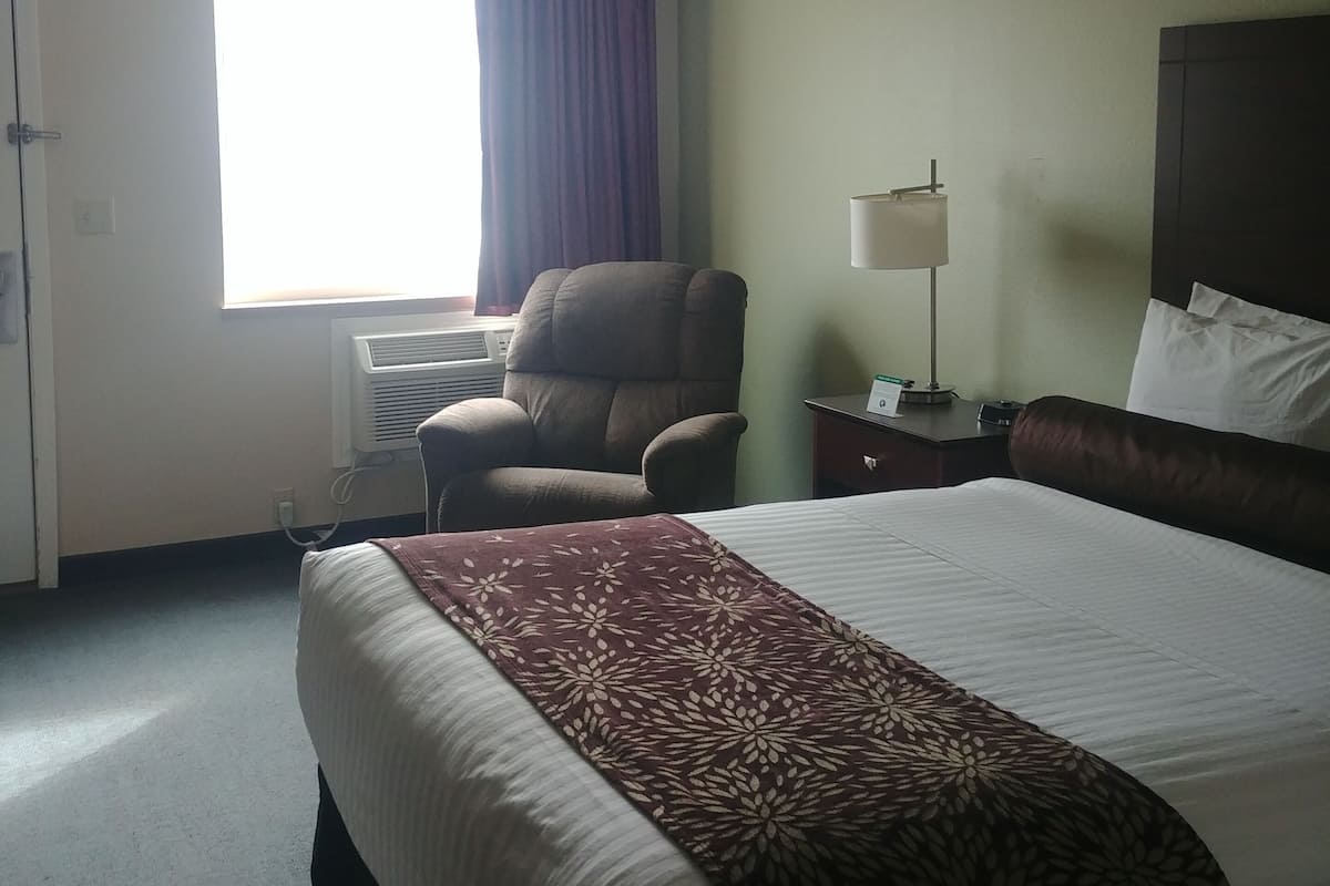 Une chambre d’hôtel avec un lit, un bureau, une chaise, une lampe et une fenêtre avec des rideaux.