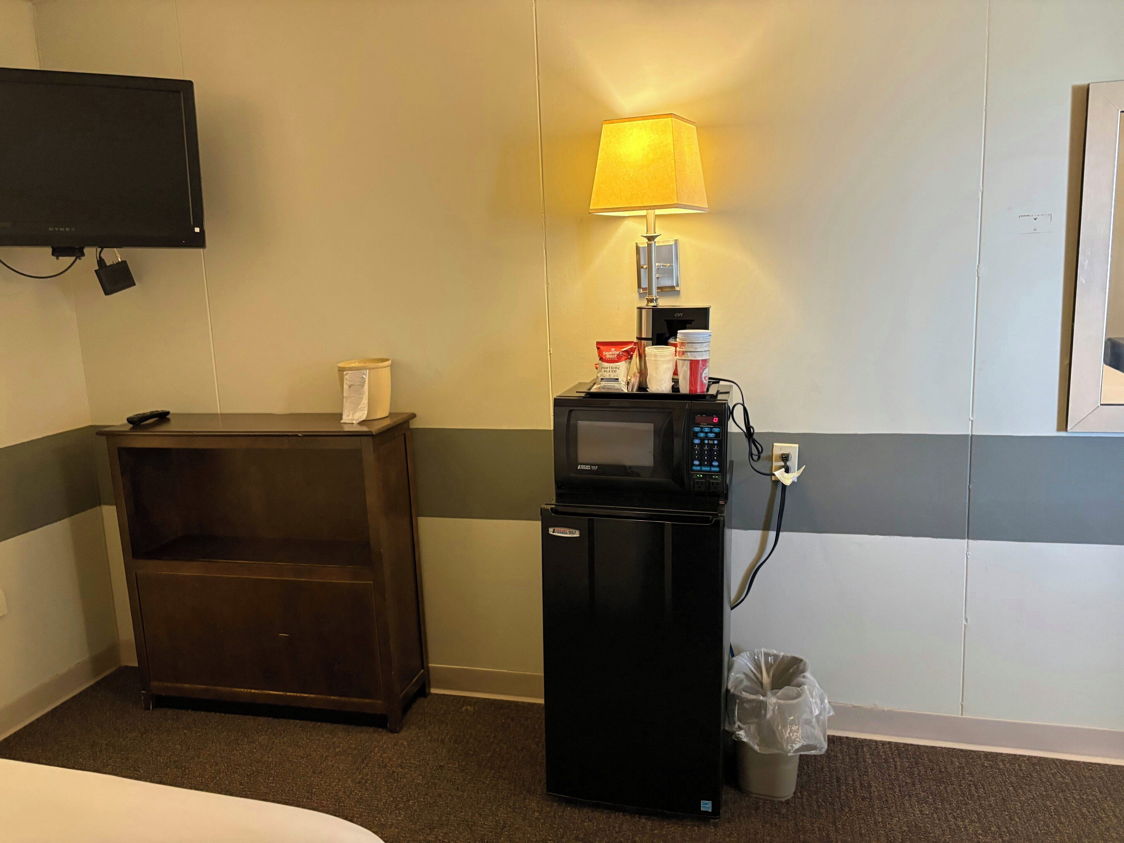 Ein Hotelzimmer mit einem Bett, einem dunklen Nachttisch aus Holz, einem schwarzen Kühlschrank, einer Mikrowelle, einem Mülleimer, einem an der Wand befestigten Fernseher und einer Lampe.