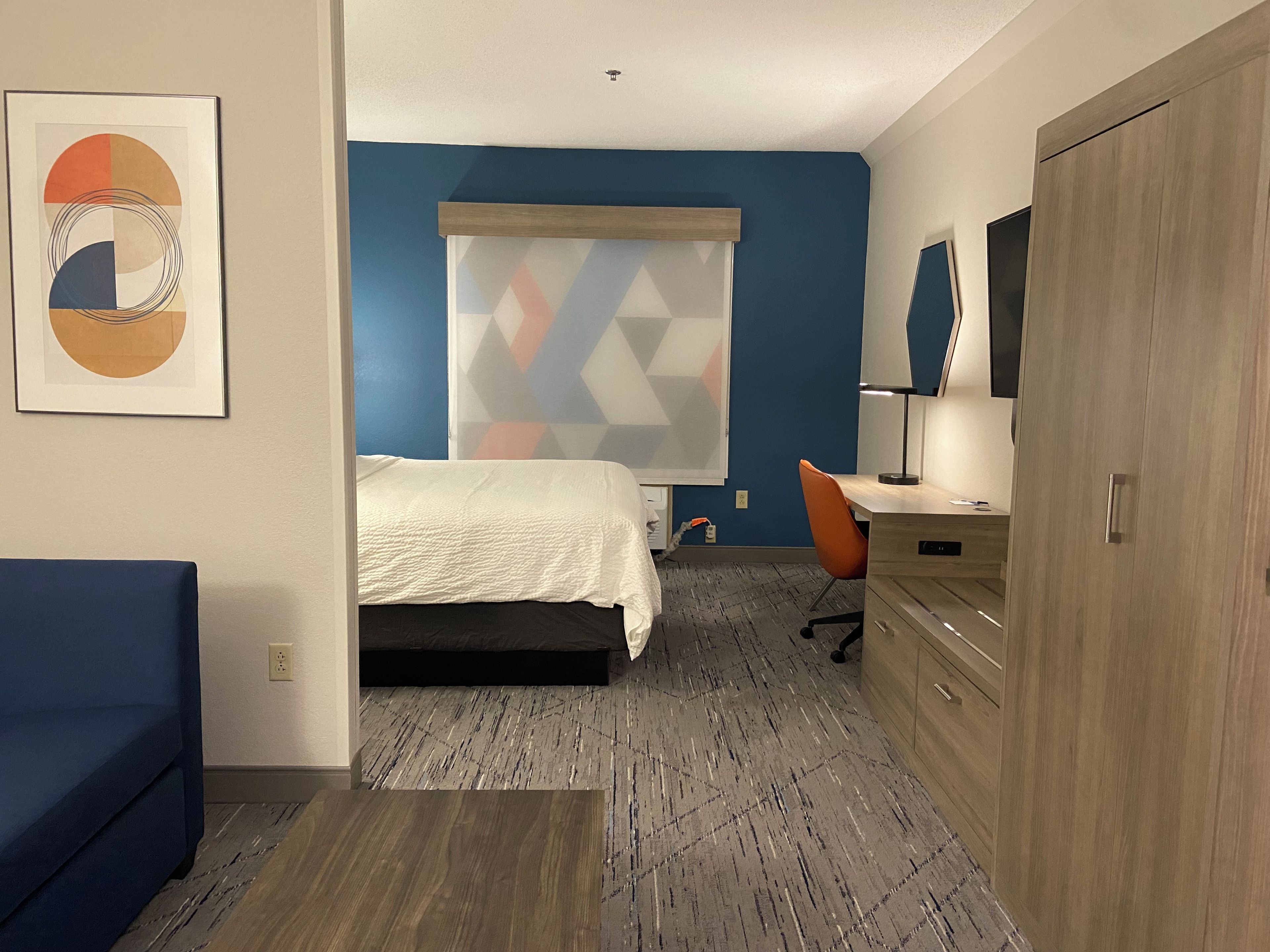 A hotel room with a bed, a desk, a chair, a wardrobe, and a wall with a geometric-patterned window blind.