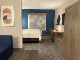 Una habitación de hotel con cama, escritorio, silla, armario y ventana con persiana de diseño geométrico.