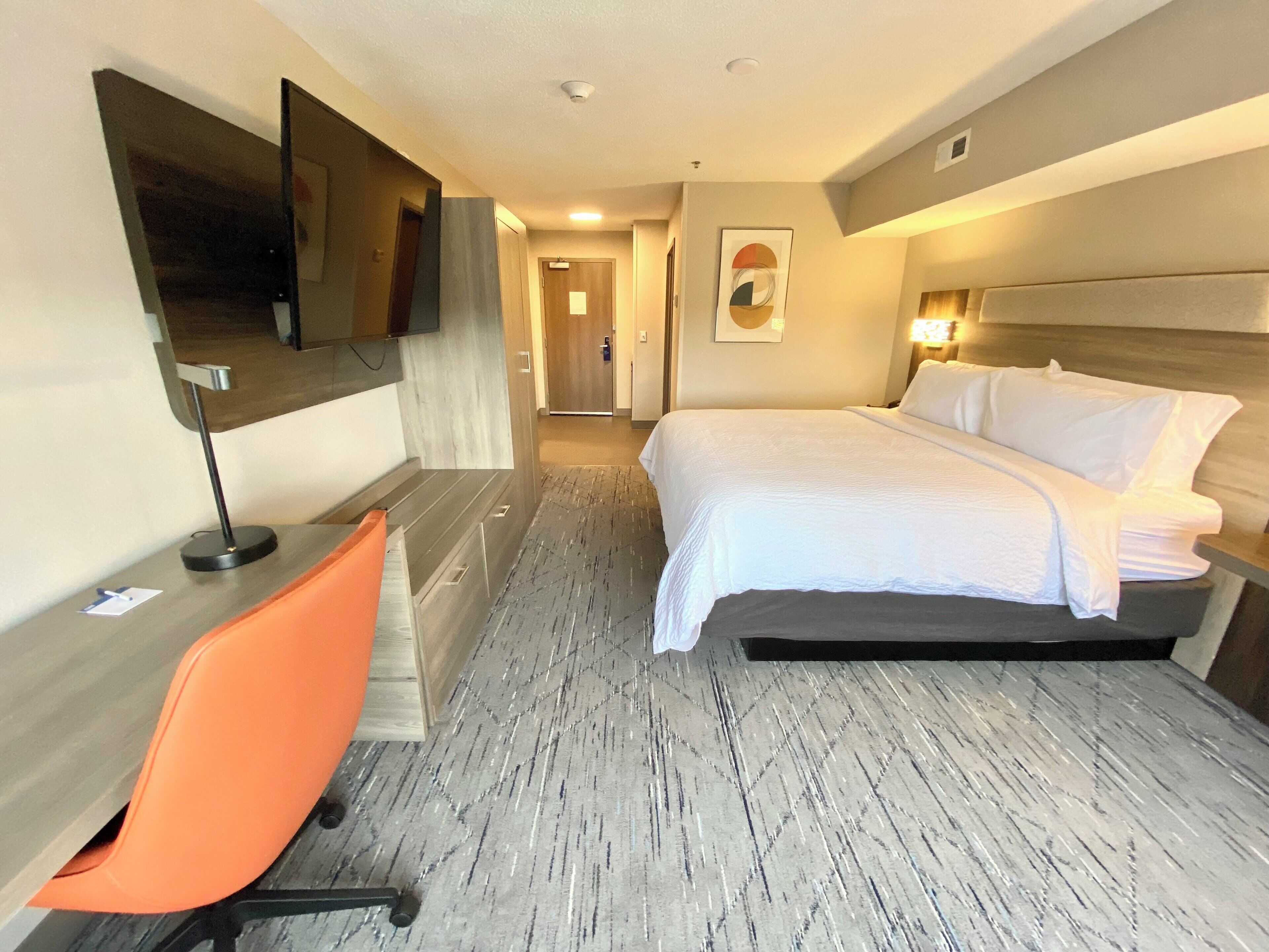 A hotel room with a bed, a desk, an orange chair, a flat-screen TV mounted on the wall, and a door leading to a hallway.