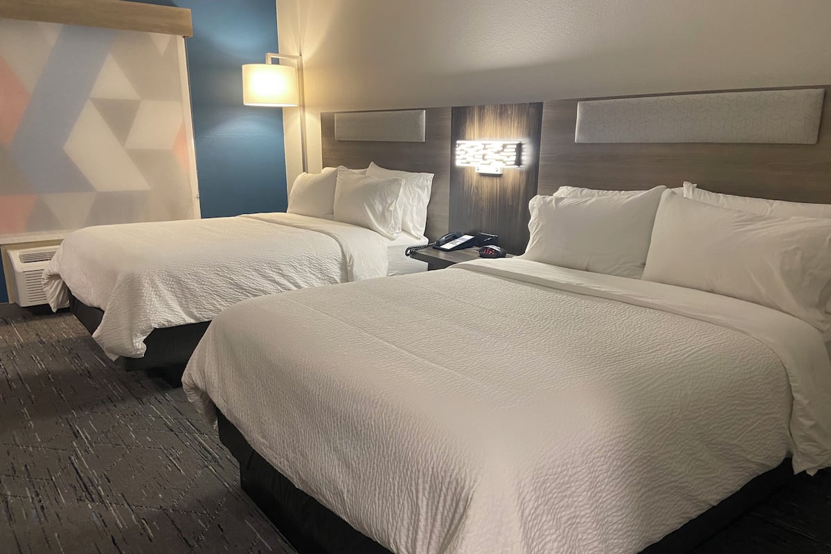 A hotel room with two beds, a bedside table with a phone and alarm clock, and a wall-mounted light fixture.