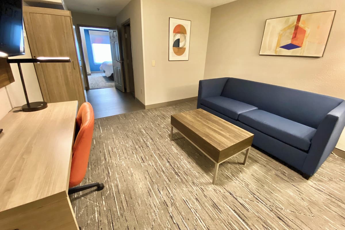 A modern living room with a blue sofa, a wooden coffee table, and a desk with a chair. There is a hallway leading to a bedroom.