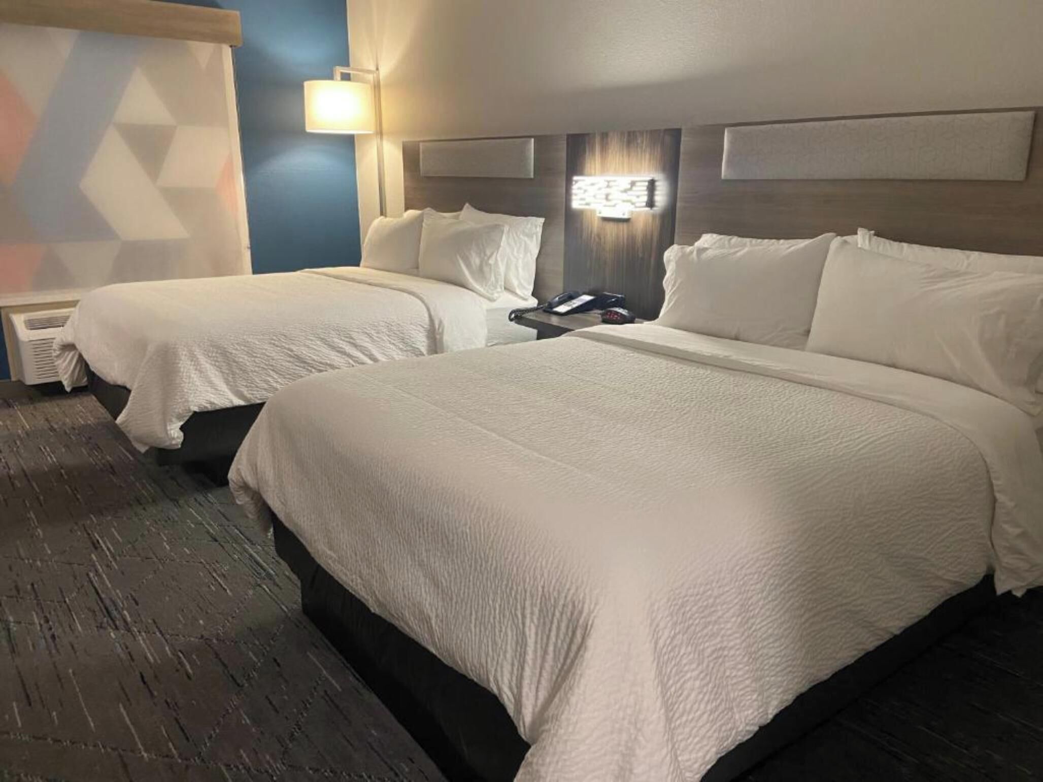 Ein Hotelzimmer mit zwei Betten, einem Nachttisch, einer Lampe und einem an der Wand hängenden Kunstwerk.