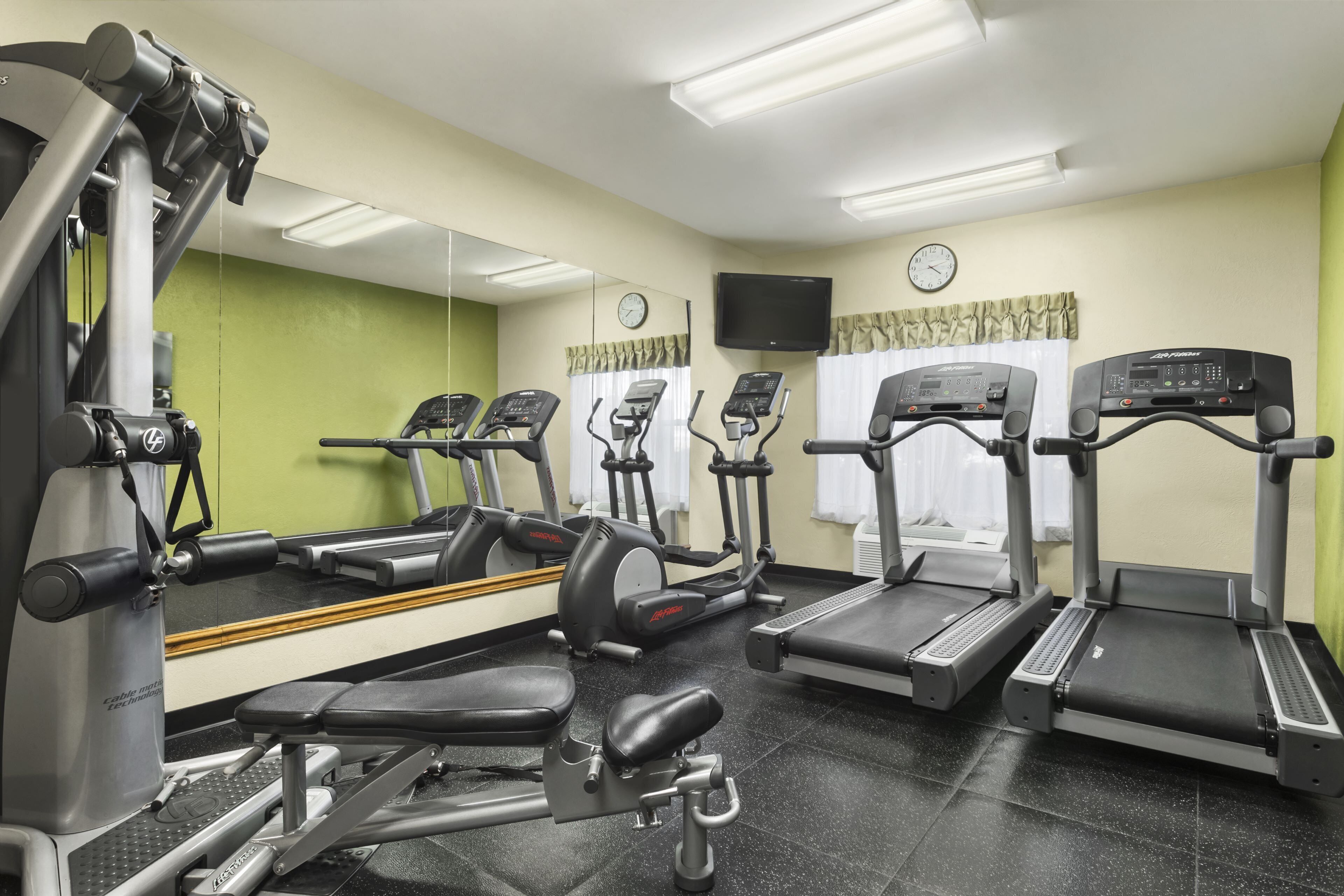 A gym with various exercise equipment, a large mirror, a clock, and a TV on the wall.