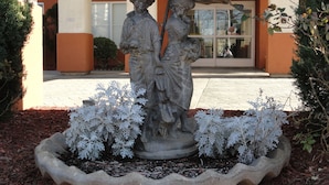 A statue of a man and woman under an umbrella, surrounded by white ornamental plants, in front of a building with orange and beige walls. - Concord Suites (Concord)