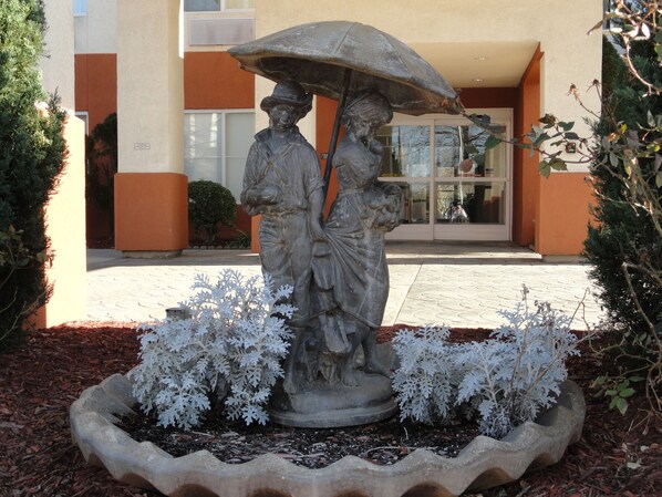 A statue of a man and woman under an umbrella, surrounded by white ornamental plants, in front of a building with orange and beige walls. - Concord Suites (Concord)
