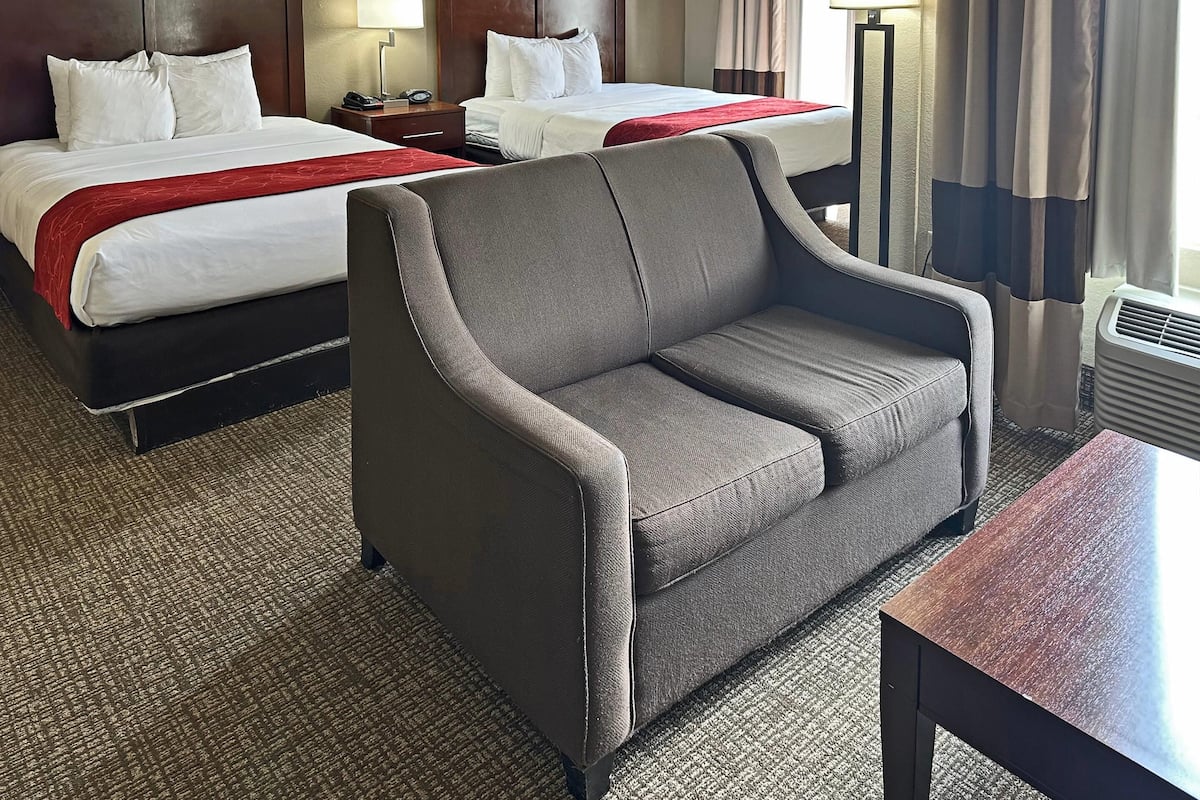 A hotel room with two beds, a grey armchair, a small table, and a lamp.