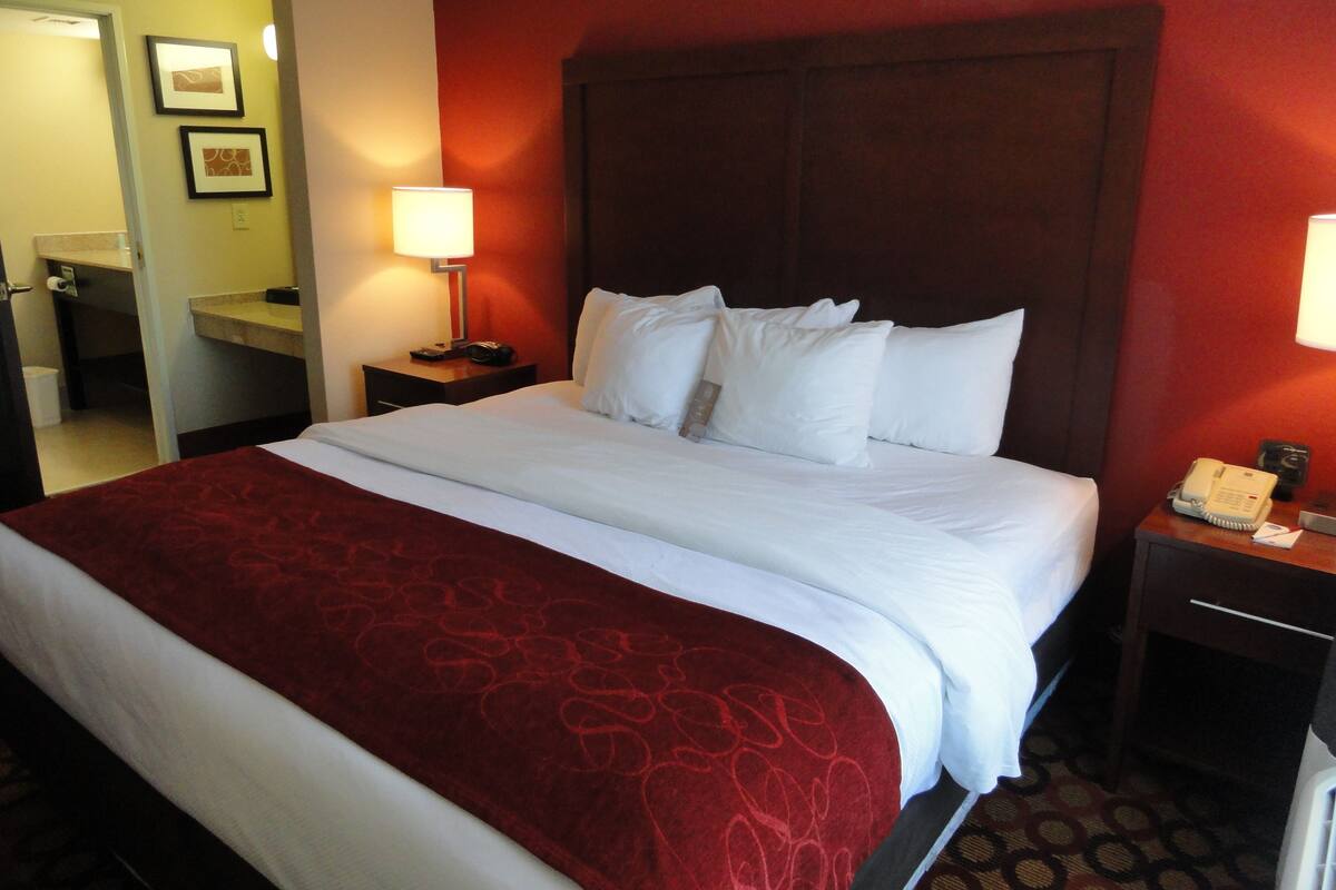 A hotel room with a large bed, two bedside tables, a nightstand, a mirror, and a bathroom visible through an open door.