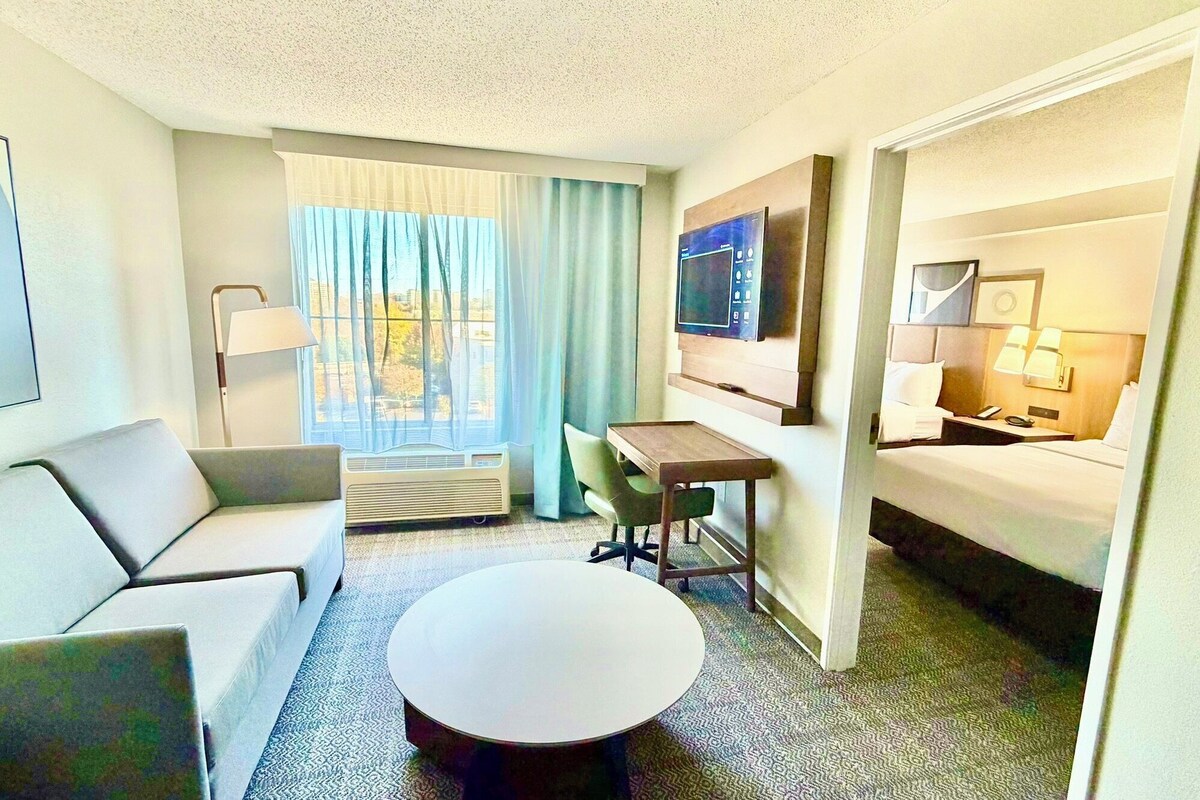 A hotel room with a sofa, a desk, a bed, a TV, and a window with curtains.