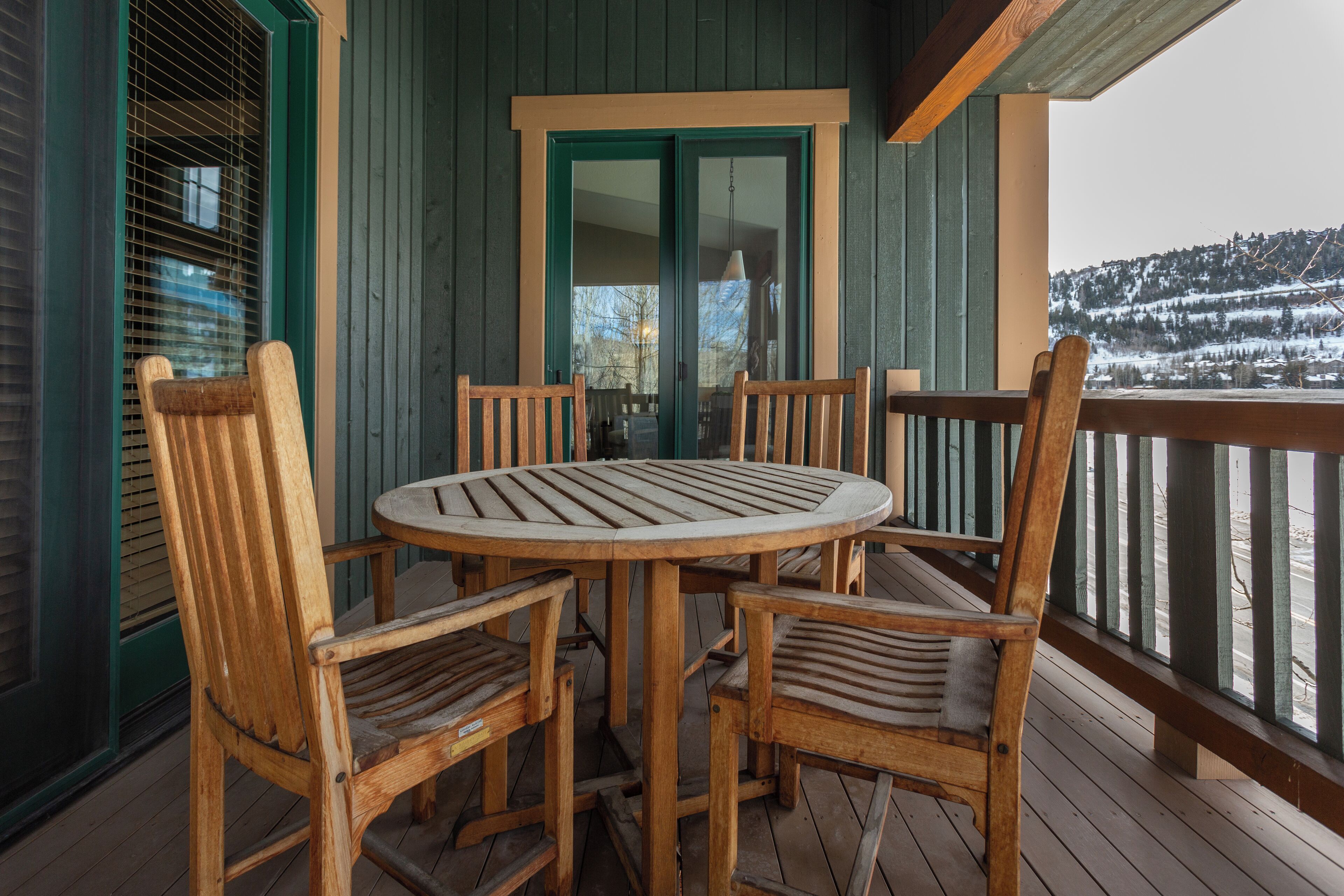 A wooden deck with a round table and chairs, a door with a green frame, and a view of a snowy landscape.