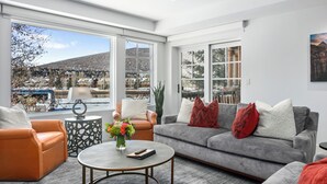 Uma sala de estar moderna com um sofá cinzento, uma poltrona laranja e uma mesa de centro redonda. Grandes janelas proporcionam vista para uma paisagem nevada e edifícios.