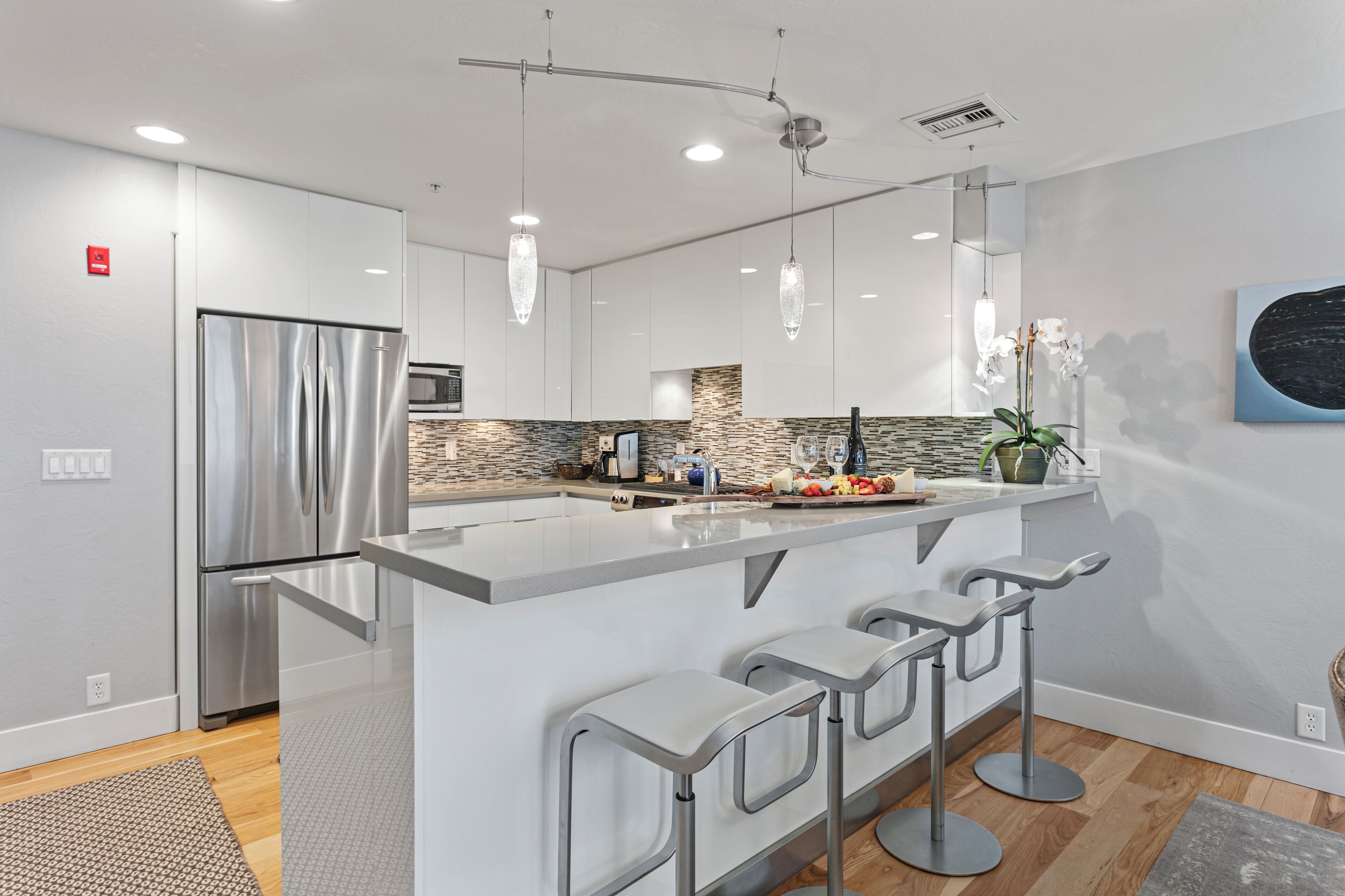 A modern kitchen with a stainless steel refrigerator, a large island with bar stools, pendant lighting, and a backsplash with stone tiles.