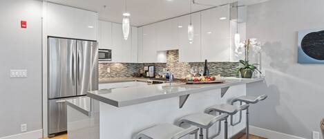 A modern kitchen with a stainless steel refrigerator, a large island with bar stools, pendant lighting, and a backsplash with stone tiles.