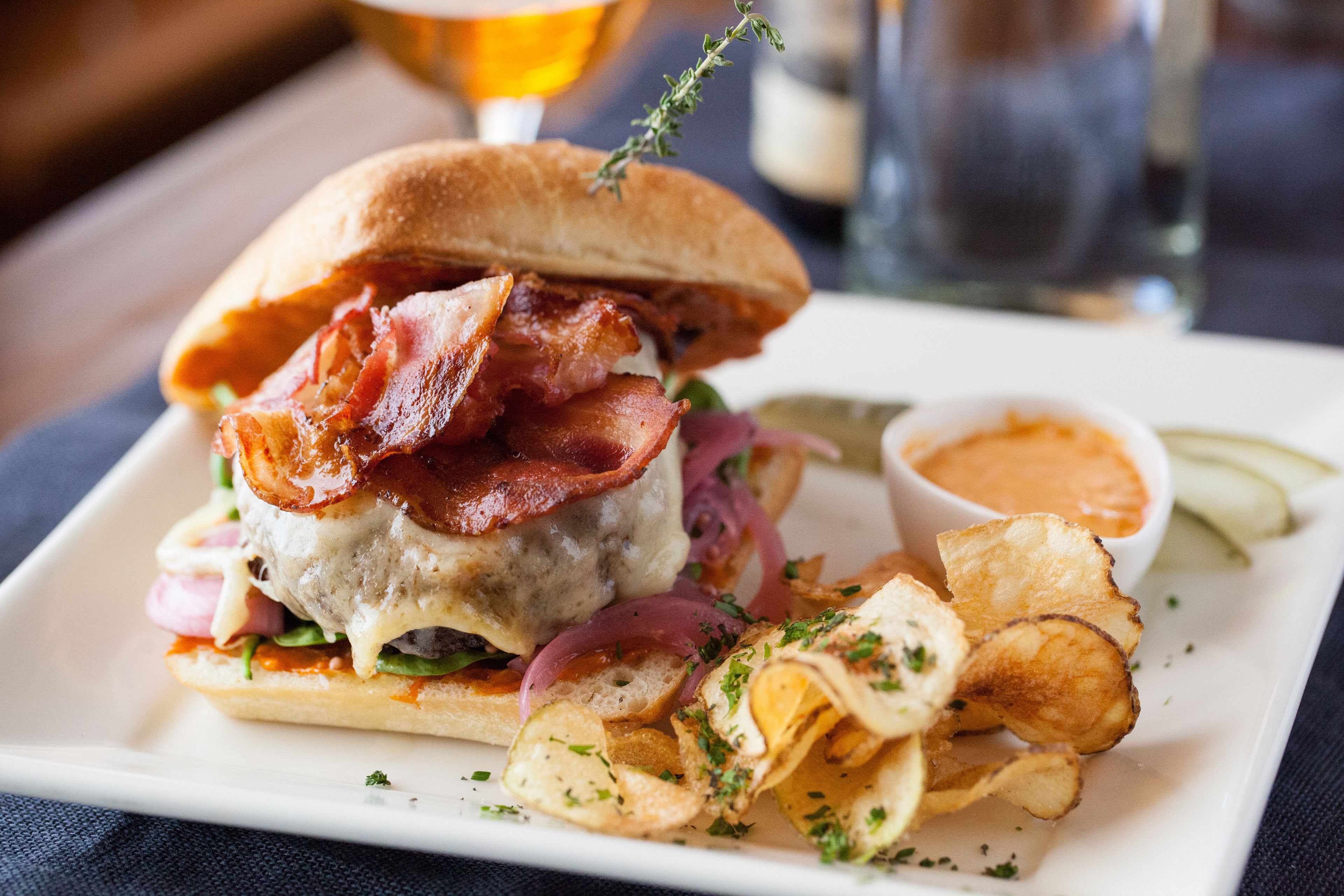 Un sándwich con tocino, una porción de papas fritas y un recipiente pequeño de salsa sobre un plato blanco.