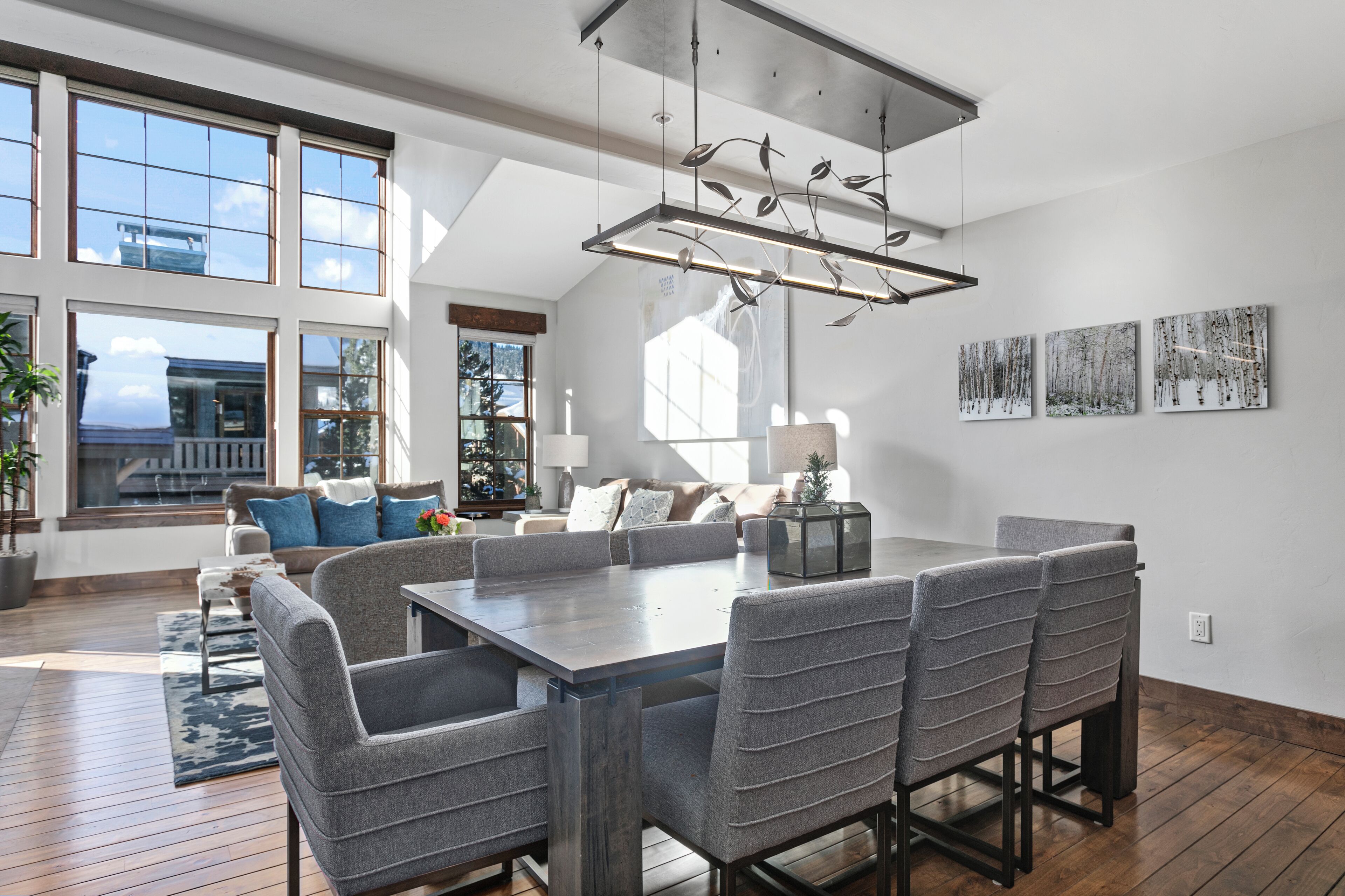 A spacious dining area with a large table and multiple chairs, a modern chandelier, and large windows offering a view of the outside.
