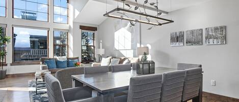 Un comedor amplio con una gran mesa y varias sillas, una lámpara moderna de araña y ventanas grandes que ofrecen vistas al exterior.