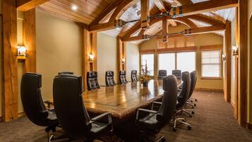 A conference room with a long wooden table surrounded by black chairs, wooden beams on the ceiling, and windows with blinds.