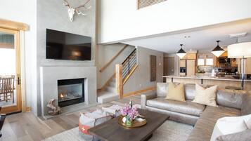 A modern living room with a fireplace, a large flat-screen TV, a sofa set, and a kitchen area in the background.