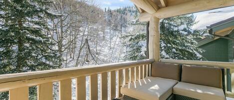 Una terraza de madera con una zona de estar acolchada y una mesa con cubierta de vidrio, con vistas a un paisaje nevado y árboles.