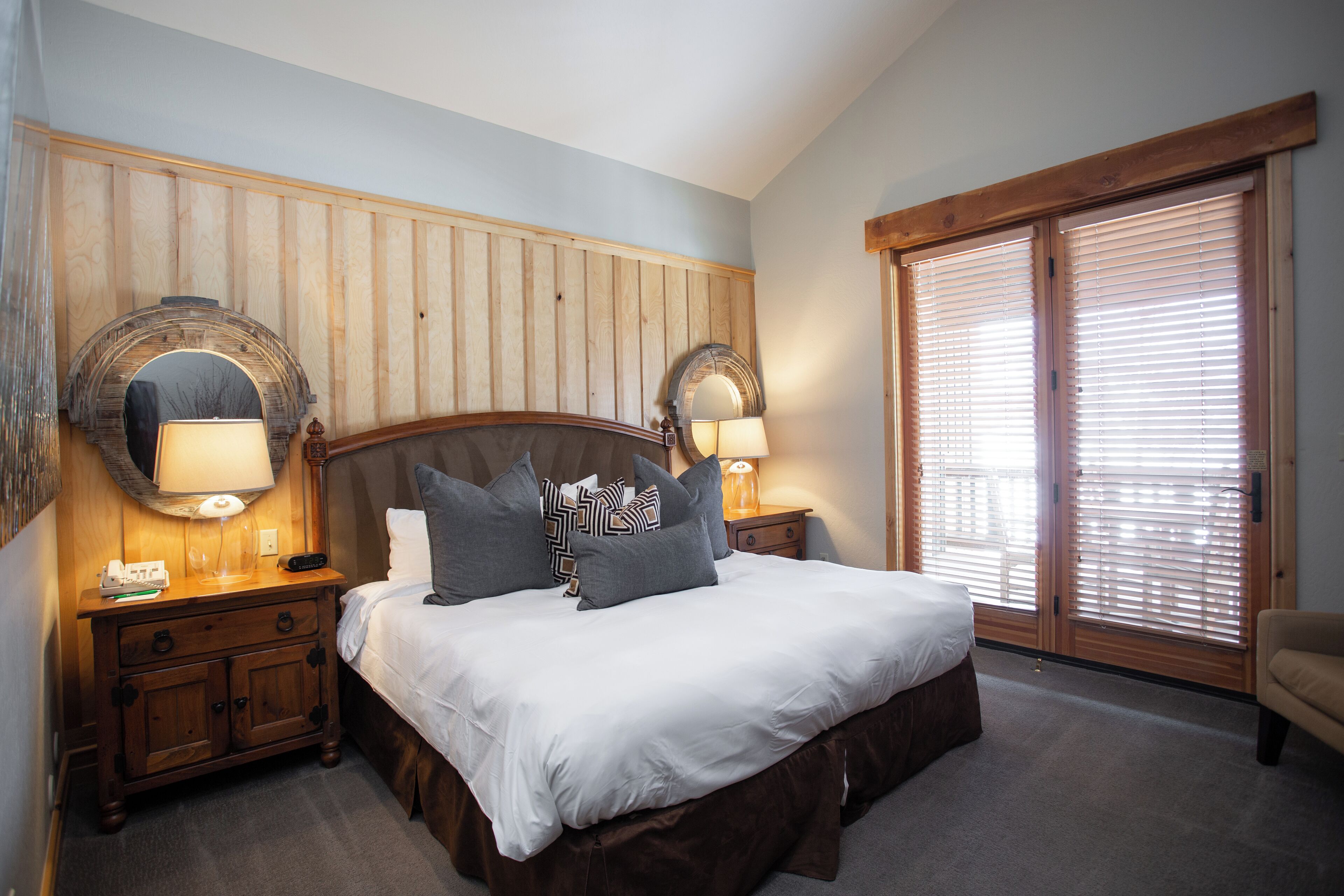 A bedroom with a large bed, wooden headboard, two bedside lamps, and a large window with blinds.