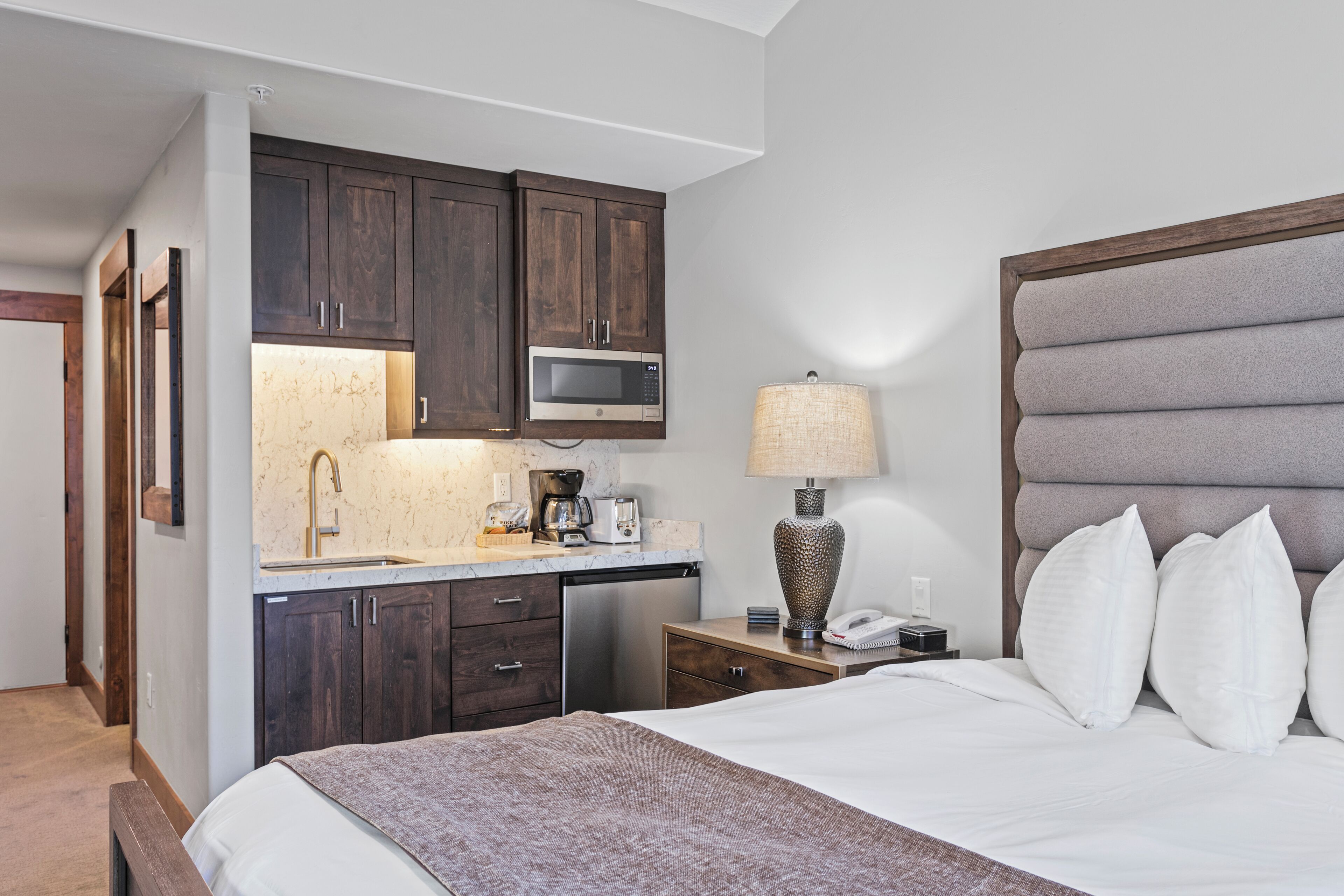 A hotel room with a bed, a nightstand with a lamp, a microwave, a sink, and wooden cabinets.