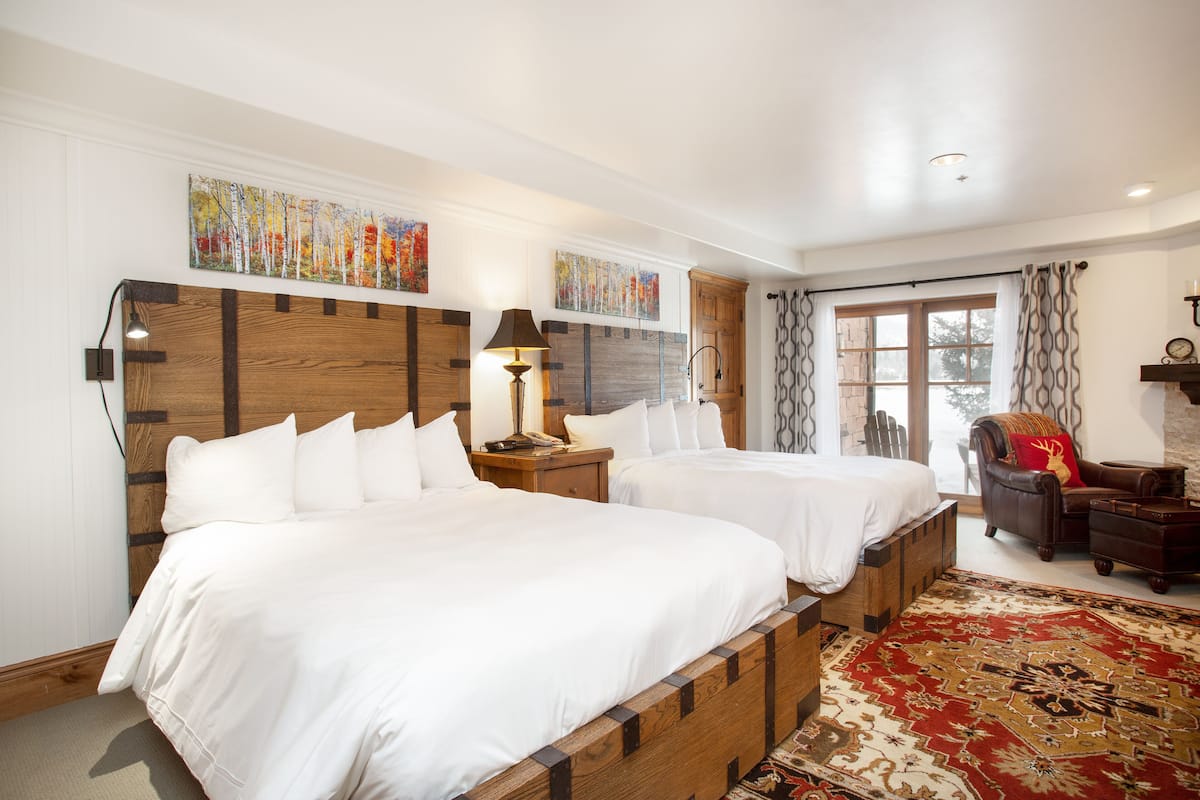 A hotel room with two beds, a wooden headboard, a red patterned rug, a leather armchair, and artwork on the walls.