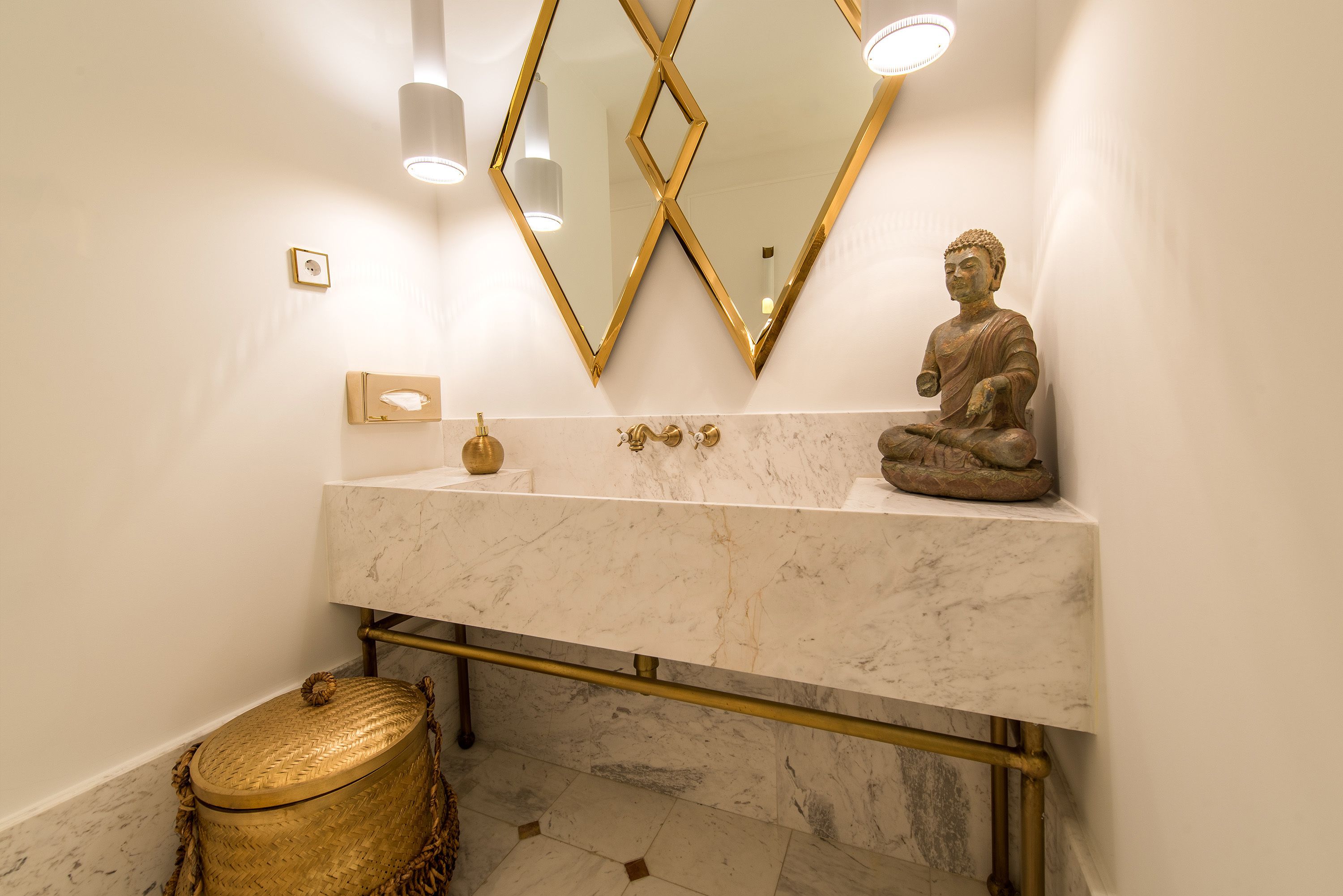 Un baño con lavamanos de mármol, detalles dorados, una estatua de Buda y una cesta tejida.