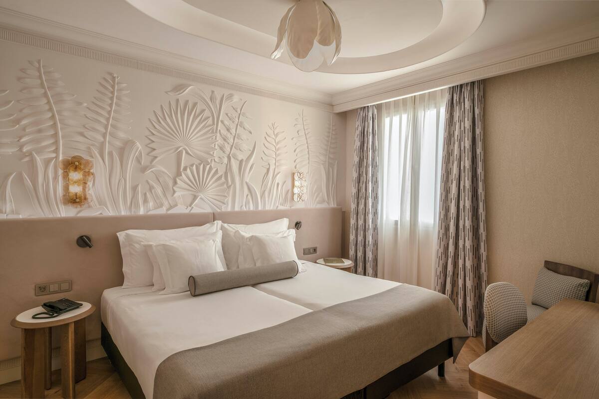 A hotel room with a large bed, a bedside table with a phone, a small table with a chair, and a decorative wall with leaf patterns.