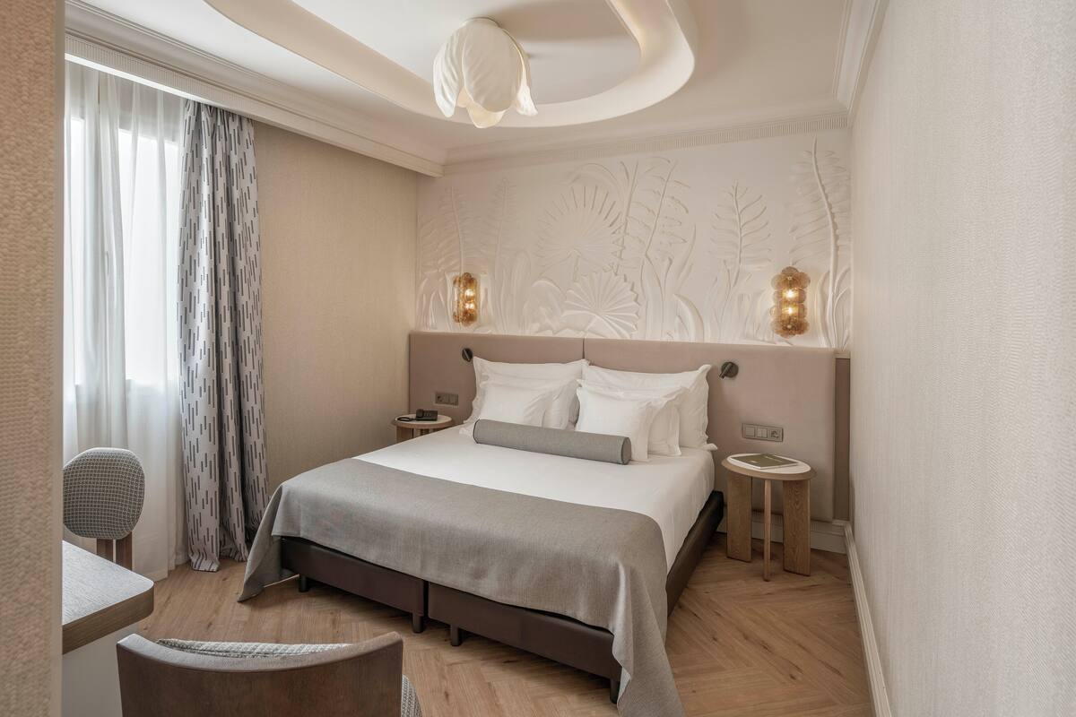 A modern hotel room with a large bed, bedside tables, a small round table, and a chair.